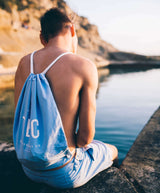 A young man sitting by the water wearing Tailored Swim Short - Sea with a drawstring bag in soft blue.